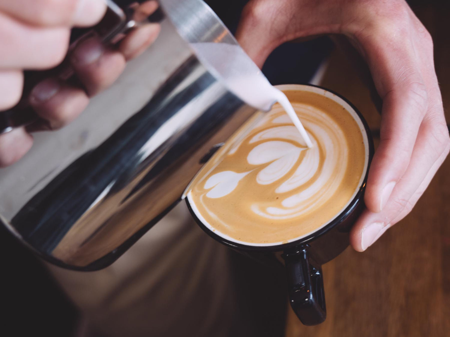 Person pouring milk into a coffee cup to create latte art on a wooden surface.